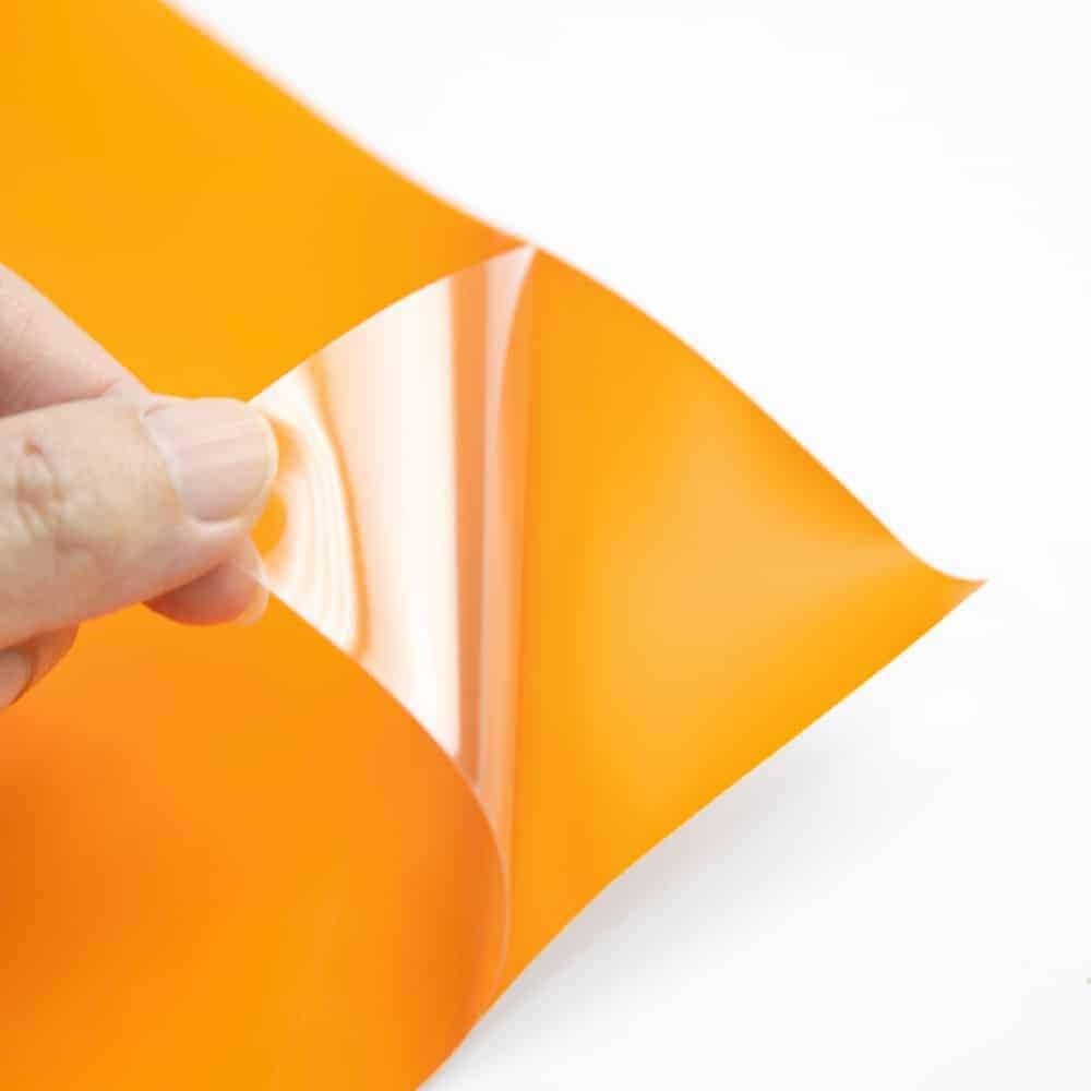 Mano levantando papel adhesivo brillante naranja para mostrar su cara adhesiva en pliego de 70x100 cm